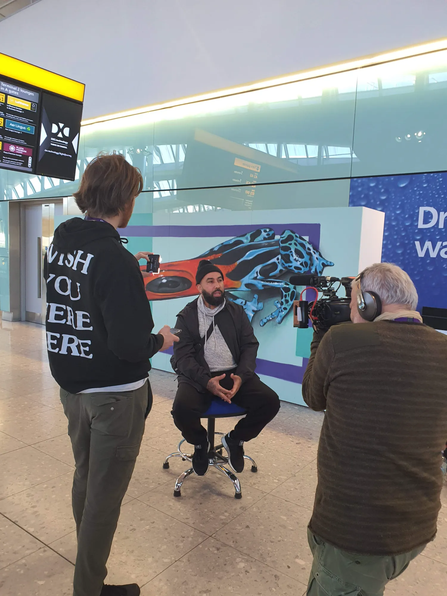 Artist Sérgio Odeith posing next to his finished 3D anamorphic blue frog mural inside London Heathrow Airport.