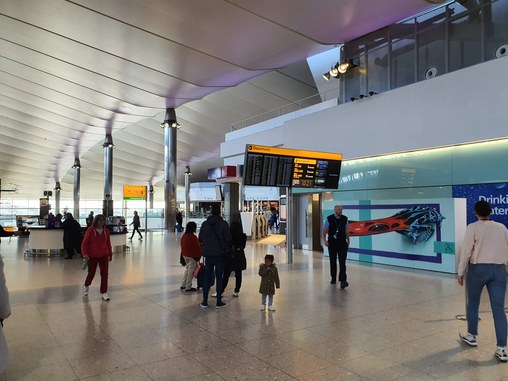 Wide shot of the Heathrow Airport departure hall showing international travelers walking past and viewing Odeith's 3D frog mural.